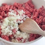Raw ground meat and chopped onions sizzle in a white pan with a wooden spatula, setting the stage for a delicious taco rice skillet.
