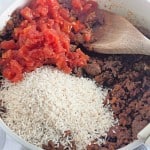 A white skillet brimming with cooked ground meat, diced tomatoes, and uncooked rice offers the perfect base for a delightful taco rice skillet. A wooden spoon casually rests on the edge, inviting you to stir up this flavorful fusion.