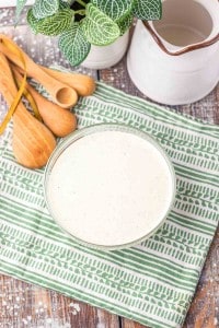 A bowl of creamy white dressing sits on a green and white patterned cloth next to wooden measuring spoons and a white pitcher, with a small green plant in the background.