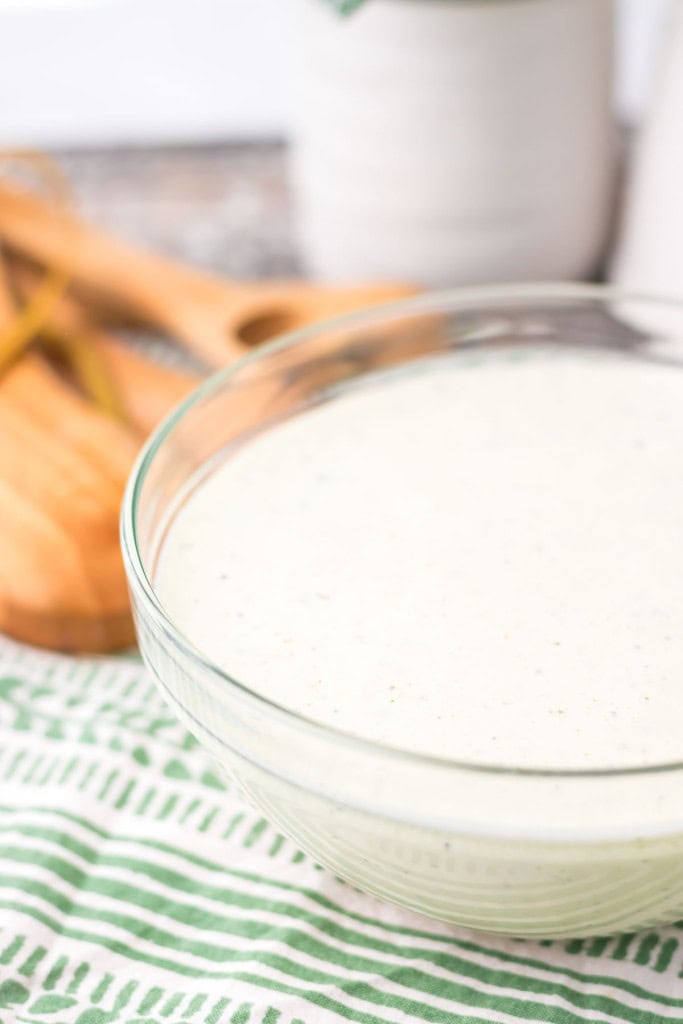 A clear glass bowl filled with a creamy white sauce sits on a green and white striped cloth. Two wooden spoons are in the background.
