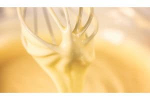 Close-up of a whisk with creamy batter drizzling off the tines against a blurred background.