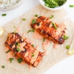 Grilled salmon fillets with sauce and chopped green onions on parchment, garnished with lemon wedges. Perfect for fish recipes, this dish is complemented by bowls of rice and extra green onions in the background.