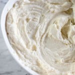 A bowl of thick creamy frosting with a metal spatula rests on a marble surface, perfect for topping those freshly baked Blueberry Danishes.
