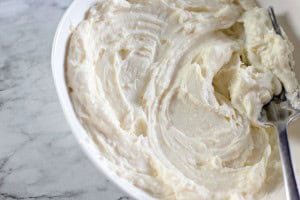 A bowl of thick creamy frosting with a metal spatula rests on a marble surface, perfect for topping those freshly baked Blueberry Danishes.