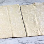 Two sheets of rolled-out puff pastry are laid on a marble surface, each with a visible fold line down the center, ready to be transformed into delicious blueberry Danishes.