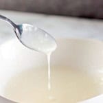 A spoon drips liquid into a white bowl on a marble surface, reminiscent of the glaze dripping from freshly baked blueberry Danishes.