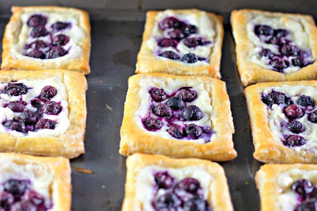 Nine rectangular pastries with a golden crust, topped with cream cheese and blueberries, resembling classic blueberry Danishes, are neatly arranged in rows on a baking sheet.