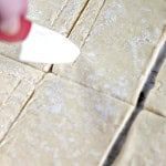 A hand with a red knife scores rectangular pieces of dough on a floured surface, preparing for delicious blueberry Danishes.