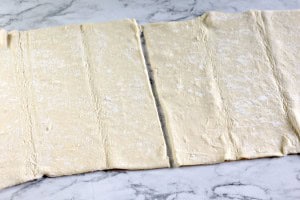 Two sheets of rolled-out puff pastry are laid on a marble surface, each with a visible fold line down the center, ready to be transformed into delicious blueberry Danishes.