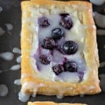 A rectangular pastry, known as a Blueberry Danish, topped with creamy icing and vibrant blueberries, rests elegantly on a dark surface.