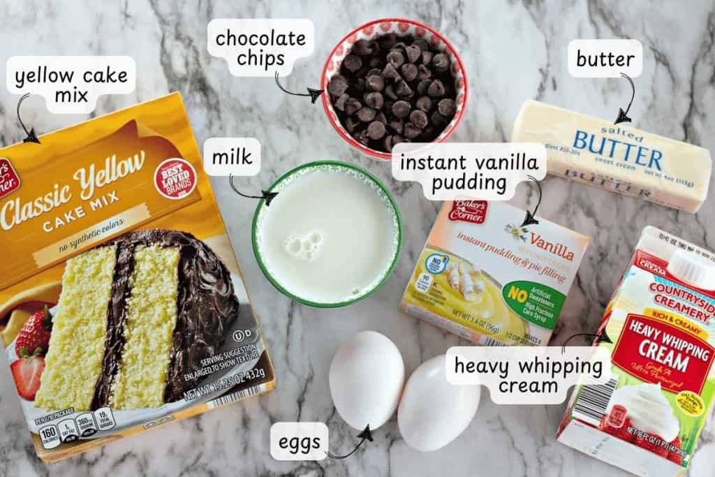 Ingredients for baking Boston Cream Pie Cookies: yellow cake mix, chocolate chips, milk, instant vanilla pudding, butter, heavy whipping cream, and two eggs, all arranged elegantly on a marble surface.
