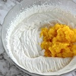 A bowl of whipped cream mixed with a mound of mashed yellow fruit rests on a marble countertop, reminiscent of the creamy texture found in a Boston Cream Pie Cookie.