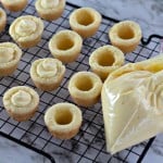 Mini tart shells with creamy filling rest on a cooling rack beside a piping bag of yellow cream, reminiscent of a Boston Cream Pie Cookie, all set elegantly on a marble counter.