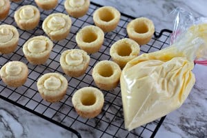 Mini tart shells with creamy filling rest on a cooling rack beside a piping bag of yellow cream, reminiscent of a Boston Cream Pie Cookie, all set elegantly on a marble counter.