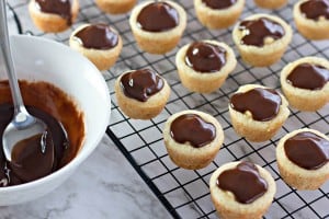 Mini tarts with chocolate filling rest on a wire rack, reminiscent of Boston Cream Pie's rich allure, next to a bowl of melted chocolate and a waiting spoon.