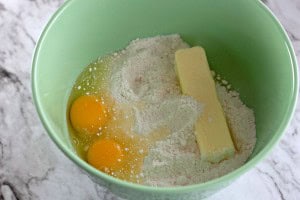 A green bowl contains two raw eggs, flour, and a stick of butter on a marble surface—the perfect start to whipping up a batch of Boston Cream Pie Cookies.