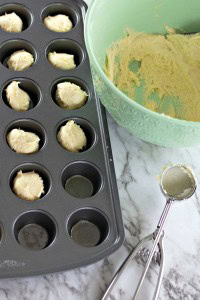 Cookie dough scoops sit in a mini muffin pan beside a green mixing bowl, with a nod to the indulgence of Boston Cream Pie Cookies, and a dough scoop rests on the marble countertop.