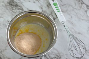 A mixing bowl with powder and liquid ingredients, preparing to create a delightful Boston Cream Pie Cookie, sits next to a metal whisk on a marble countertop.