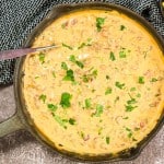 A skillet filled with creamy cheese dip topped with chopped green herbs. A spoon rests in the skillet.