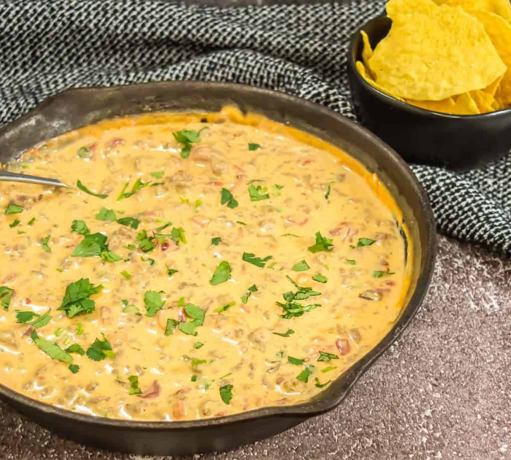 A skillet of creamy cheese dip with herbs on top, placed on a textured surface. A small black bowl of tortilla chips is beside it.