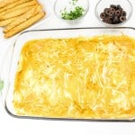 A glass dish of baked enchiladas topped with melted cheese, surrounded by cilantro, black olives, and rolled tortillas on a white background.