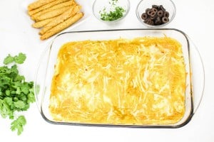 A glass dish of baked enchiladas topped with melted cheese, surrounded by cilantro, black olives, and rolled tortillas on a white background.