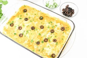 Casserole dish topped with melted cheese, sliced black olives, and chopped cilantro. Two bowls on the side contain sour cream and more sliced olives.