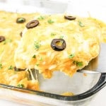 A slice of cheesy casserole with olives and herbs is being served from a glass baking dish.