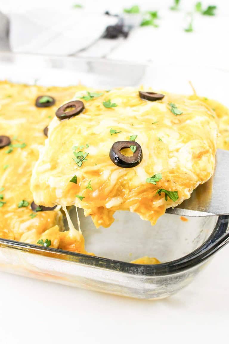 A slice of cheesy casserole with olives and herbs is being served from a glass baking dish.