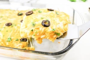 A slice of cheesy enchilada casserole with black olives and herbs is lifted from a glass baking dish.