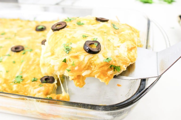 A slice of cheesy enchilada casserole with black olives and herbs is lifted from a glass baking dish.