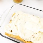 A glass baking dish with a layer of cake covered in white frosting. A spatula rests on top. Nearby containers hold various ingredients.