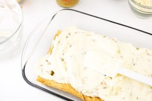 A glass baking dish with a layer of cake covered in white frosting. A spatula rests on top. Nearby containers hold various ingredients.