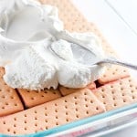 A delightful Strawberry Ice Cream Cake features layered biscuits and strawberry filling in a glass dish, topped with whipped cream being spread with a spoon.