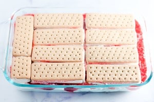A glass baking dish showcases a strawberry ice cream cake-inspired layered dessert: vanilla wafer cookies on top, encasing a layer of red gelatin, with fluffy whipped topping visible underneath.