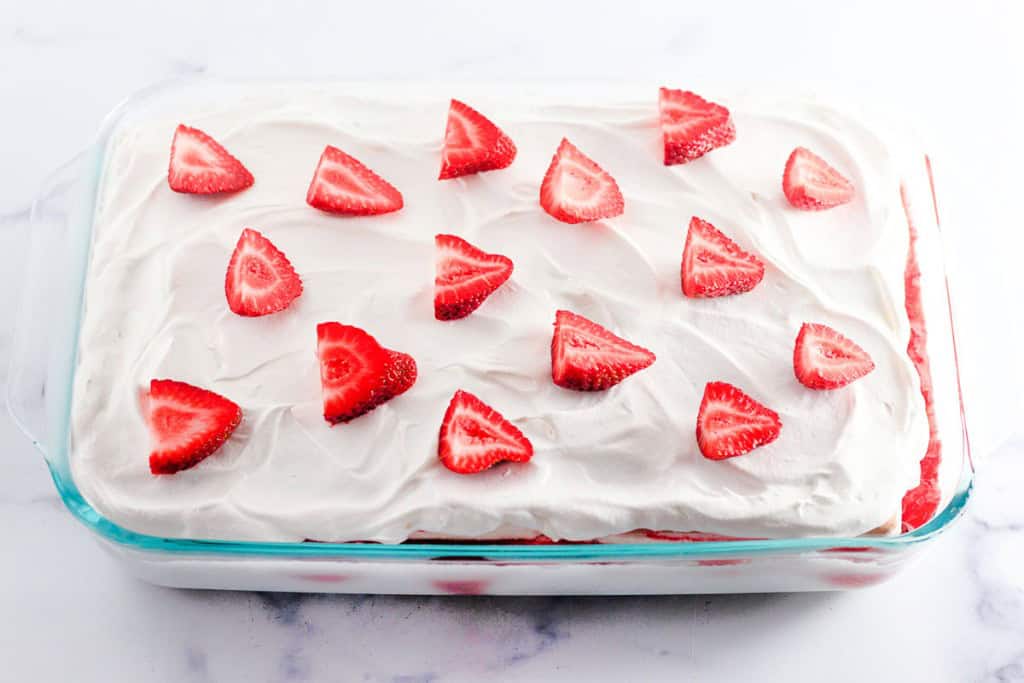 This delightful strawberry ice cream cake features a rectangular shape, topped with white whipped delight and artfully decorated with evenly spaced strawberry slices, all presented in a clear glass dish.