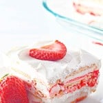 Square slice of layered strawberry ice cream cake with whipped cream and a strawberry slice on top, served on a white plate next to a whole strawberry.