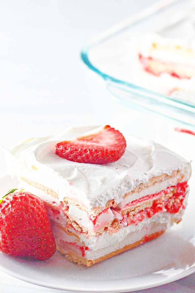 Square slice of layered strawberry ice cream cake with whipped cream and a strawberry slice on top, served on a white plate next to a whole strawberry.