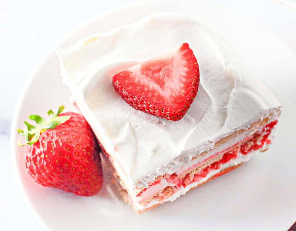 A slice of strawberry ice cream cake with white frosting, crowned with a heart-shaped strawberry slice, sits next to a whole strawberry on a white plate.