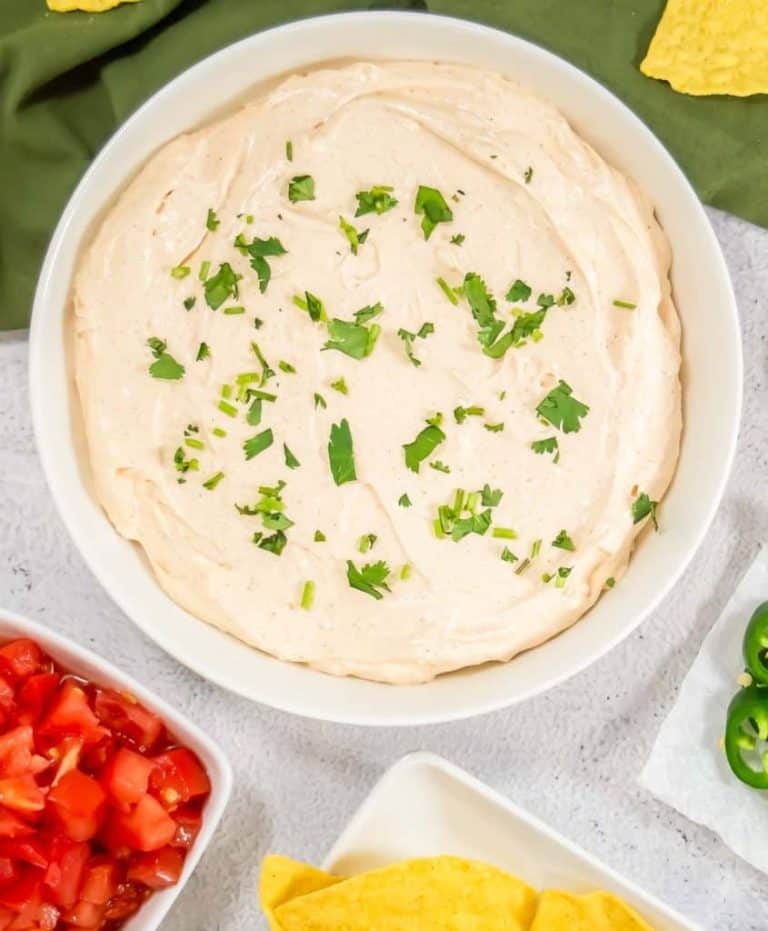 A bowl of creamy dip topped with chopped herbs, surrounded by diced tomatoes, tortilla chips, and sliced jalapeños.