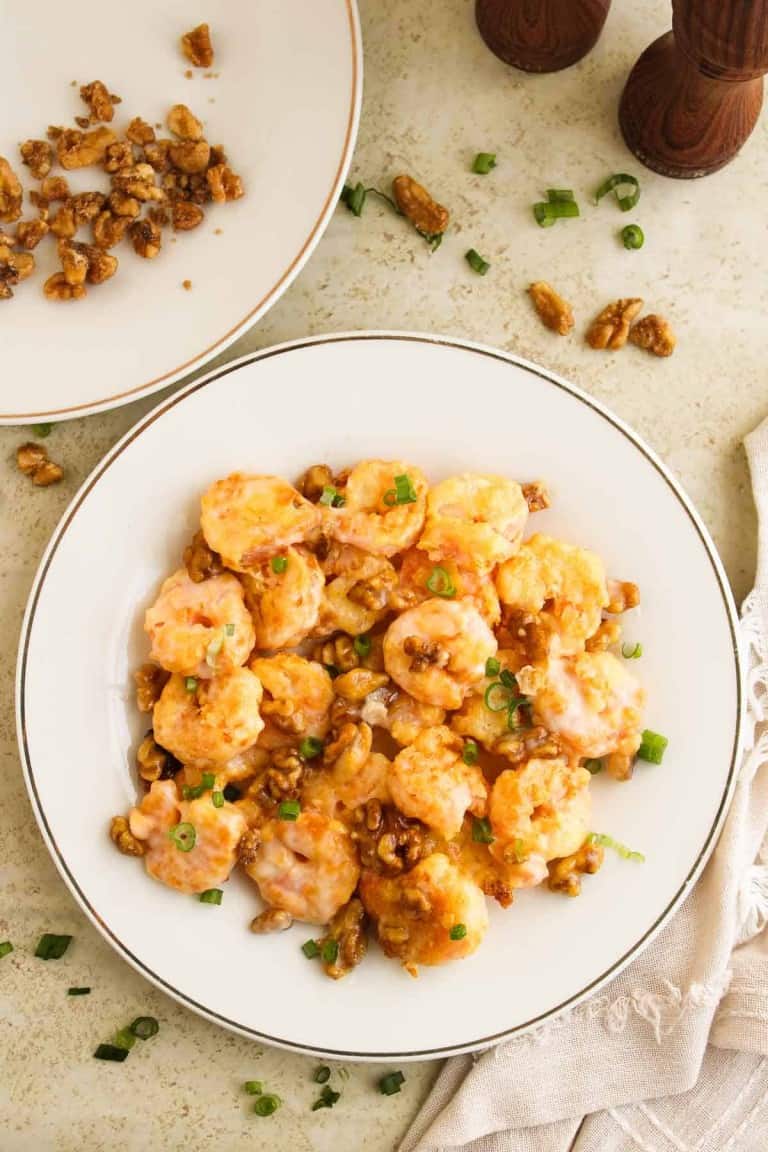 A plate of honey walnut shrimp garnished with chopped green onions, next to a napkin and pepper grinders. A separate dish of walnuts is partially visible.