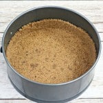 A metal springform pan containing a crumbly graham cracker crust sits on a wooden surface.