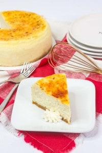 A slice of cheesecake with a caramelized top on a white square plate, garnished with whipped cream, sits on a red checkered cloth. A whole cheesecake, plates, and cutlery are in the background.
