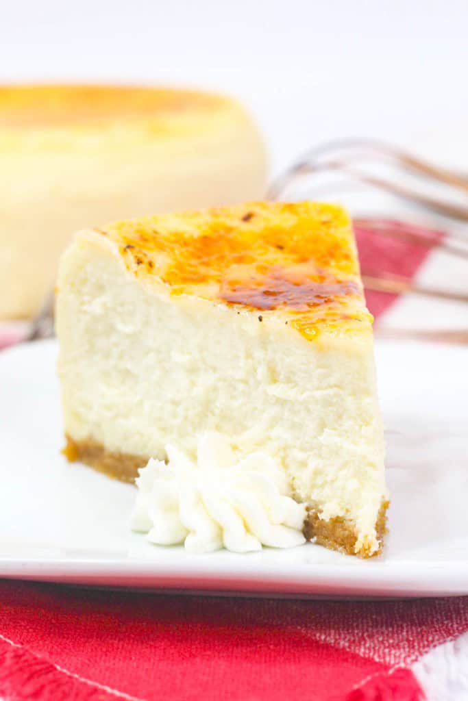 A slice of cheesecake with a caramelized top sits on a white plate, garnished with a dollop of whipped cream, on a red and white cloth background.