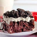 A slice of chocolate cake on a plate, topped with whipped cream and crushed chocolate cookies, next to a fork and a checkered red napkin.