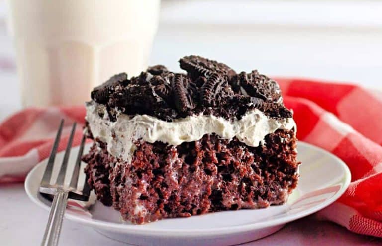 A slice of chocolate cake on a plate, topped with whipped cream and crushed chocolate cookies, next to a fork and a checkered red napkin.
