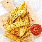 Crispy potato wedges topped with herbs and cheese on a wooden board, served with a side of ketchup.