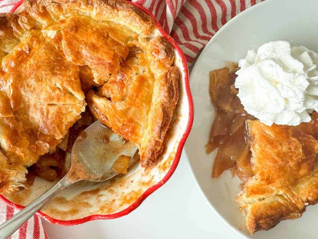 A pie dish with a partially served apple pie and a metal serving spoon. A plate next to it holds a slice of pie topped with whipped cream. A red-and-white striped cloth is visible.