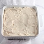 A square white baking dish filled with frosted cake rests on the marble countertop, reminiscent of Strawberry Crunch Bars.