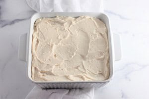A square white baking dish filled with frosted cake rests on the marble countertop, reminiscent of Strawberry Crunch Bars.
